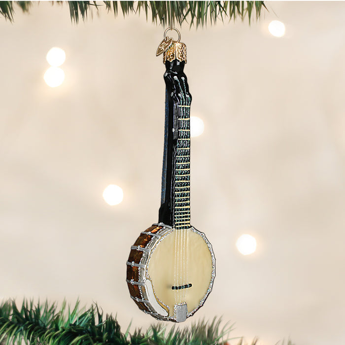 A Christmas tree ornament shaped like a banjo, featuring a cream-colored body, black neck, and silver details, hangs from a branch with blurred white lights in the background.