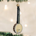 A Christmas tree ornament shaped like a banjo, featuring a cream-colored body, black neck, and silver details, hangs from a branch with blurred white lights in the background.