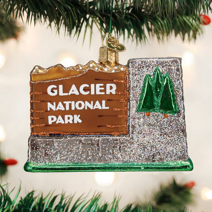 A decorative ornament shaped like the Glacier National Park entrance sign hangs on a Christmas tree, featuring glittery mountains, green trees, and white text on a brown and silver background.