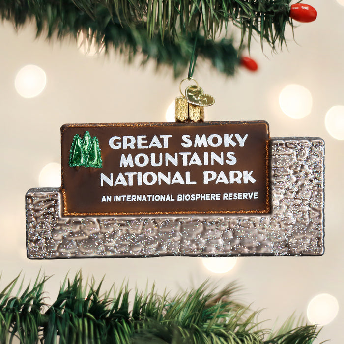 A glittery ornament shaped like the Great Smoky Mountains National Park entrance sign hangs on a Christmas tree branch, with blurred lights in the background.
