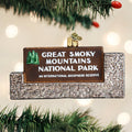 A glittery ornament shaped like the Great Smoky Mountains National Park entrance sign hangs on a Christmas tree branch, with blurred lights in the background.