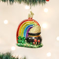 A Christmas tree ornament shaped like a rainbow ending in a pot of gold, with a green four-leaf clover and red mushrooms at the base, hanging among green pine branches with soft, glowing lights in the background.