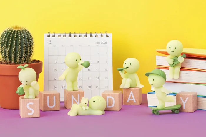 Smiski Sunday Series toys, including a singing figure, sunbathing figure, and skateboarding figure, displayed with a calendar and cactus on a table.