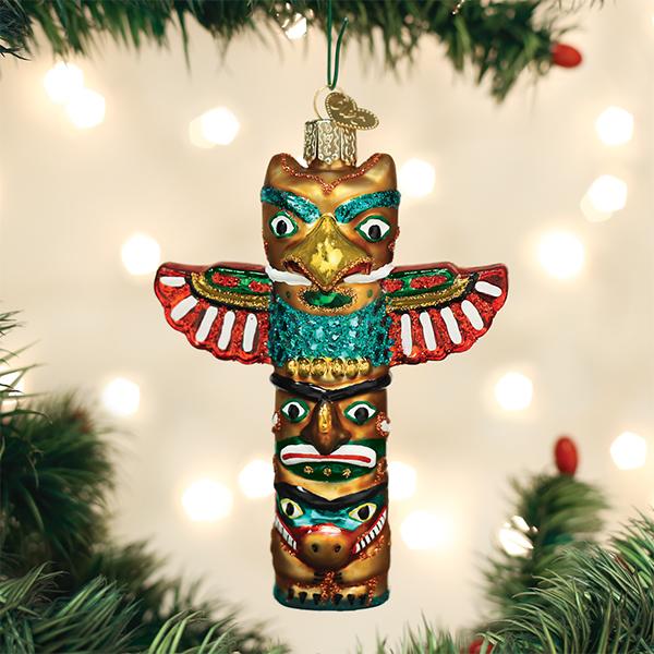 A colorful totem pole-shaped Christmas ornament hangs on a tree branch with blurred white lights in the background. The ornament features intricate designs, vibrant reds, greens, blues, and golds, and stylized faces.