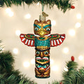 A colorful totem pole-shaped Christmas ornament hangs on a tree branch with blurred white lights in the background. The ornament features intricate designs, vibrant reds, greens, blues, and golds, and stylized faces.