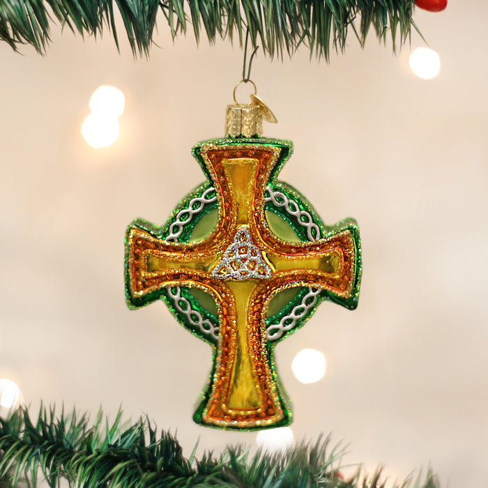 A green and gold cross-shaped Christmas ornament with intricate beading hangs on a Christmas tree branch, with blurred white holiday lights in the background.