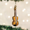 A shiny violin-shaped Christmas ornament hangs from a green tree branch, with soft white lights blurred in the background.